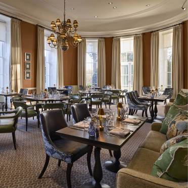 Elegant restaurant interior with round tables set for dining, upholstered chairs, a chandelier, and large windows with cream curtains. Patterned carpet and dark wood furniture are present.