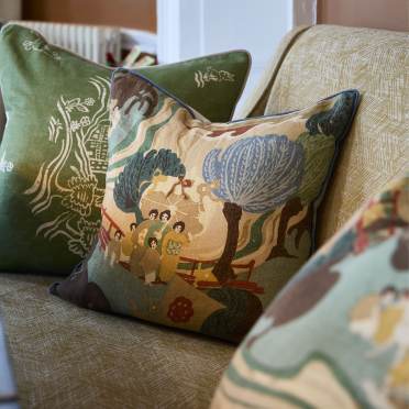 Decorative cushions with detailed, colorful patterns on a textured beige sofa. One cushion features an abstract nature and figure scene, while others have nature motifs.