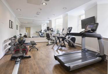 Gym A gym with treadmills, stationary bikes, a rowing machine, free weights, and benches on a wooden floor. Mirrors and a wall-mounted TV are also visible.