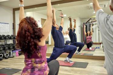 Fitness classes Four people in workout clothes perform yoga poses on mats in a gym with mirrored walls and gym equipment in the background.