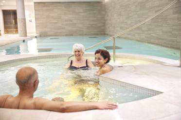 Three people relaxing in a hot tub at an indoor pool facility. Two women are seated in the water, facing a man who's sitting on the tub's edge.