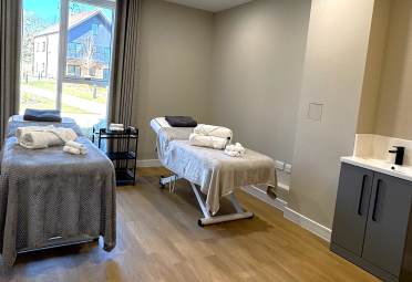 Two massage tables with gray blankets and towels in a well-lit room, featuring a window with a view outside, a cart with supplies, and a sink with a cabinet.