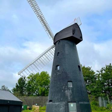 A black-painted windmill with white blades stands in front of a fence and green trees under a partly cloudy sky.