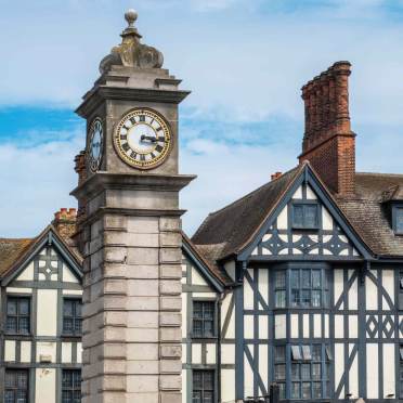 A stone clock tower stands in front of a timber-framed building with steep roofs and multiple chimneys under a partly cloudy sky.