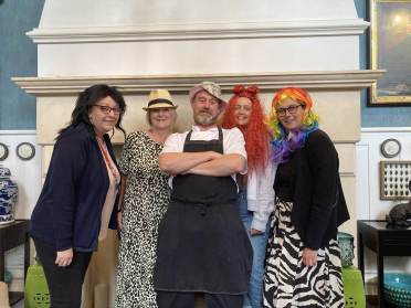 Five adults stand indoors in front of a fireplace, wearing various costumes and colorful wigs, posing and smiling for the camera.