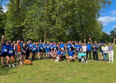 A large group of people, some with dogs, stand on grass in front of trees, wearing athletic clothes and race bibs, smiling and waving at the camera.