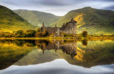 Golden hour in Argyll and Bute (Scotland) Ancient stone castle ruins stand by a calm lake, reflecting in the water, with green hills and cloudy sky in the background.