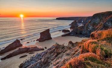 Golden hour in Cornwall Sunset over a rocky coastline with steep cliffs, sandy beach, and wildflowers in the foreground. Waves gently reach the shore.