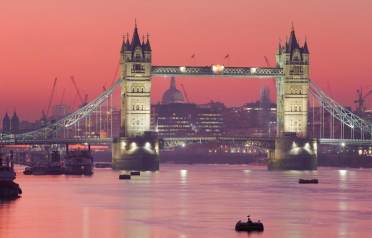 Golden hour in London Tower Bridge in London is illuminated at dusk, with calm water in the foreground and the city skyline visible under a pink-orange sky.