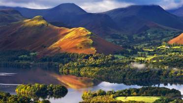 A sunlit hill with red and green slopes reflects in a calm lake, surrounded by trees and distant mountains under a partly cloudy sky.