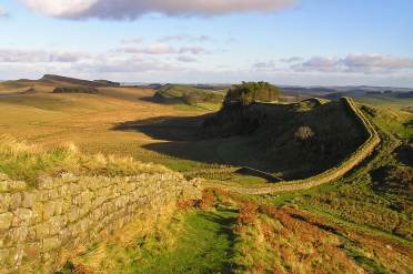 Golden hour - North East (England) A stone wall winds across a grassy, hilly landscape under a blue sky with scattered clouds. Shadows from the hills and trees are visible on the ground.