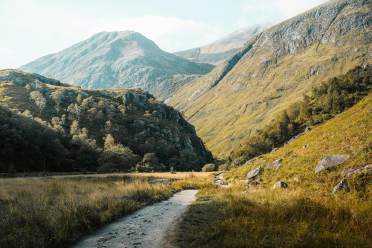 A dirt path winds through a grassy valley with steep, rocky mountains in the background under a clear sky.