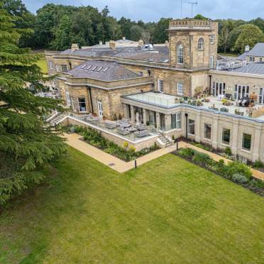 A large, historic stone building with a tower sits amid green lawns and landscaped gardens, featuring outdoor seating areas and surrounded by trees.