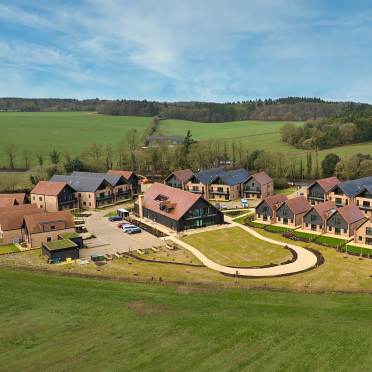 Aerial view of a modern housing complex with multiple buildings, green lawns, a curved driveway, parked cars, and surrounding countryside.