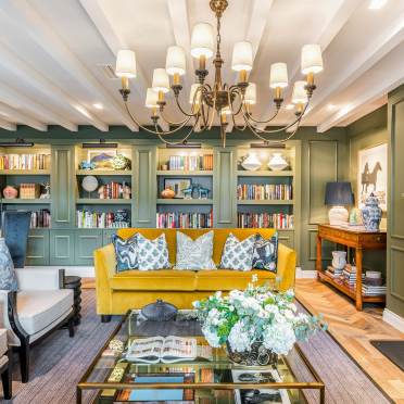 A cozy living room with a yellow sofa, patterned pillows, built-in bookshelves, a chandelier, glass coffee table, and floral arrangement.
