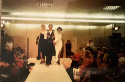 Three people walking down a runway in formal attire during a fashion show, with an audience seated on both sides.