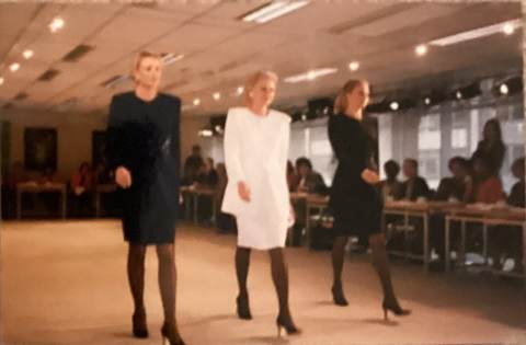 Three women in professional attire walk confidently on a runway, with an audience seated along the sides.