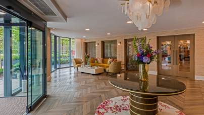 Modern hotel lobby with herringbone wood flooring, a round table with a flower arrangement, seating area with sofas, glass doors, and decorative lighting fixture.