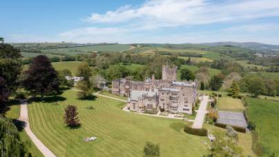 Aerial view of a large stone mansion surrounded by expansive green lawns, trees, and countryside under a clear blue sky.