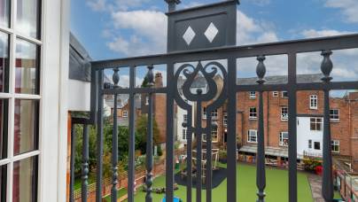 View from a balcony with ornate black railing overlooking a school playground with artificial grass and outdoor play equipment, surrounded by brick buildings under a partly cloudy sky.