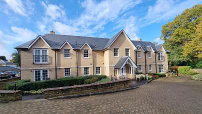 A two-story beige brick residential building with multiple gables, white trim, and small balconies, set against a blue sky with scattered clouds.