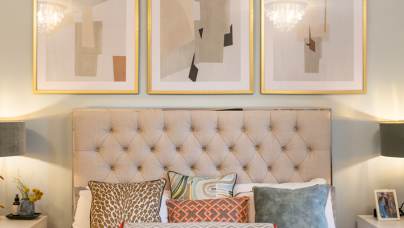 A neatly made bed with assorted pillows sits below three abstract framed artworks; two side tables hold lamps and small decorative items.