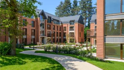 Modern brick apartment buildings with large windows and balconies, surrounded by green lawns and trees, under a clear blue sky. A curved path leads through the landscaped area.