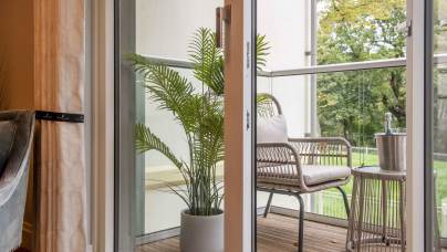 A balcony with wicker seating and a potted plant is seen through an open glass door. A small table holds a champagne bottle in an ice bucket. Trees are visible in the background.