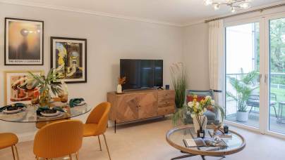 A modern living room with a round dining table, orange chairs, a TV on a wooden console, and decorative art on the walls. Large window overlooking a balcony with plants.