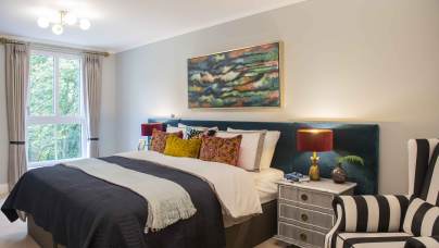 A bedroom with a large bed, colorful pillows, two nightstands with lamps, a painting above the bed, a striped armchair, and a window with curtains.