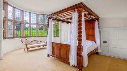 A bedroom with a wooden four-poster canopy bed, sheer white curtains, a curved vintage chaise lounge, and large bay windows overlooking a green landscape.