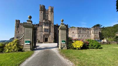 A historic stone castle with two tall towers, an arched entrance, and a gravel driveway, surrounded by manicured gardens and lawns under a clear blue sky.