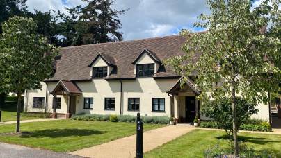 A two-story cream-colored building with a steep tiled roof, black window frames, and two entrances, surrounded by grass, trees, and a pathway under a partly cloudy sky.