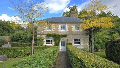A two-story stone house with a central door, surrounded by a manicured garden, featuring two small trees in front and hedges along a brick pathway.
