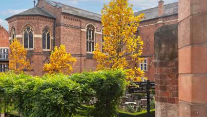 A brick church with arched windows is surrounded by vibrant autumn trees and greenery. A small signpost is visible in the garden area.