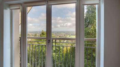 View through large glass doors of a balcony with a railing, overlooking a landscape with trees and distant fields under a partly cloudy sky.