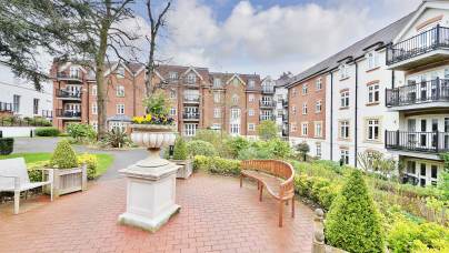 A courtyard with a brick path, benches, a large planter with flowers, and residential buildings in the background. Cloudy sky overhead.