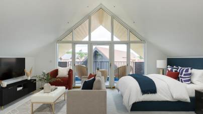 A spacious bedroom with a vaulted ceiling features a bed, a TV, a red couch, a beige loveseat, and a small balcony with outdoor chairs, all under large triangular windows.