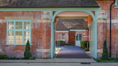 A brick building with an arched entrance leading to a courtyard. It features large windows and a pathway bordered by small plants.