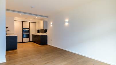 Modern kitchen with white walls, wooden flooring, and built-in appliances, viewed from an adjacent open area with recessed lighting.