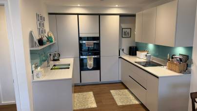 Modern kitchen with white cabinets, turquoise backsplash, wooden floor, and integrated appliances. Countertops hold a kettle, toaster, and other kitchen items. Wall shelves display decor.