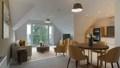 Modern attic apartment with a living area, dining table, and kitchen. Features sloped ceilings, large windows, and yellow chairs.