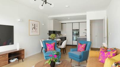 Modern living room with blue and yellow chairs, a wooden coffee table, and a wall-mounted TV. A kitchen is visible in the background, and bright cushions add color to the space.