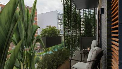 A small urban balcony with a chair, side table, and plants. Vertical trellis with greenery partially obstructs the view of neighboring buildings.