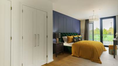 Modern bedroom with a large bed, mustard duvet, green upholstered headboard, and wooden flooring. There are white closets, a navy accent wall, and a window with a view of greenery.