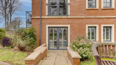 Pathway leads to a modern brick building with large windows and patio doors, surrounded by shrubs and a wooden bench in the foreground.