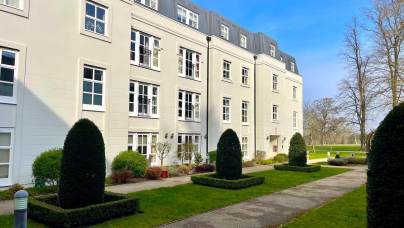 A beige multi-story residential building with dormer windows, manicured hedges, and a walkway through a grassy courtyard on a clear day.