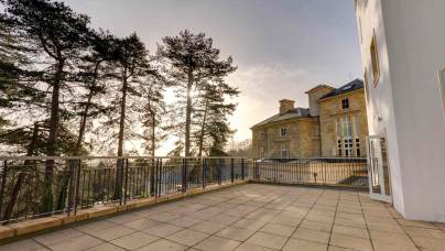 Large terrace with stone flooring and railing, adjacent to a historic building. Tall trees in the background with sunlight filtering through.