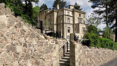 Large stone mansion with tall columns and ornate details, surrounded by lush greenery. Stone wall and stairs lead up to the entrance. Bright, partly cloudy sky in the background.