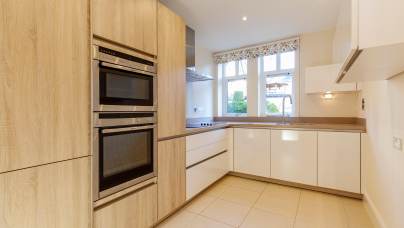 Modern kitchen with light wood cabinetry, stainless steel appliances, and white countertops. A window with floral curtains overlooks a tiled floor.